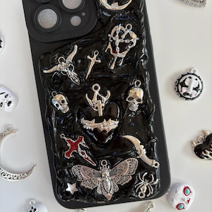 Op de afbeelding: Zwarte telefoonhoes met een textuur, versierd met zilveren bedels. Bedels zijn onder meer een schedel, kruis, mot en halve maan. De hoes heeft een zwarte cameralensbeschermer.