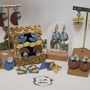 Peut inclure: Collection de boucles d'oreilles et accessoires en pâte polymère faits à la main. Les bijoux présentent des motifs bleus, jaunes, marrons et vichy. Les boucles d'oreilles sont exposées sur des présentoirs dorés et un miroir encadré, mettant en valeur le style artistique.
