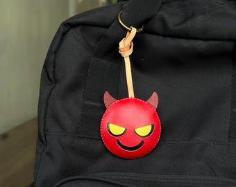 May include: A red leather devil face keychain with yellow eyes and horns, attached to a black backpack. The keychain is round and has a smiling expression. The keychain is attached to a gold-colored ring.