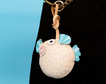 May include: A whimsical pufferfish keychain in cream-colored leather, adorned with light blue and pink accents. The fish features a stitched design and is attached to a leather loop and metal chain, perfect for adding a touch of fun to any accessory.