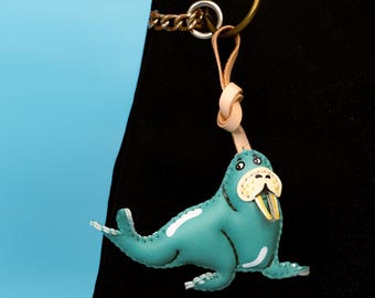 May include: A teal leather walrus keychain with white accents and a tan leather loop. The walrus has a yellow tusk and a white and tan face. The keychain is attached to a gold-colored ring and chain.