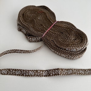 May include: A roll of brown ribbon featuring a white floral pattern along its edges. The ribbon is coiled and held together with a pink elastic band. A section of the ribbon is unrolled, displaying the intricate design.