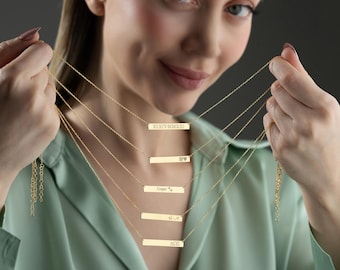 May include: Gold-colored bar necklaces with delicate chains. Each bar is engraved with text, including Roman numerals, names, and symbols. The necklaces are displayed against a soft green shirt.