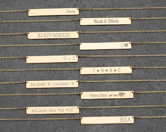 Op de afbeelding: Goudkleurige staafkettingen met gegraveerde teksten. De inscripties bevatten namen, initialen, data, coördinaten en zinnen als "Mama Bear". Elke ketting heeft een delicate ketting en een rechthoekige hanger.