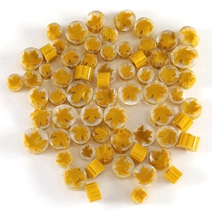 Op de afbeelding: Een verzameling gele glazen kralen met een esdoornbladontwerp. De kralen zijn helder en hebben een matte afwerking.