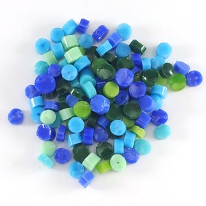May include: A collection of small, round glass beads in shades of blue, green, and turquoise. The beads are scattered on a white surface.