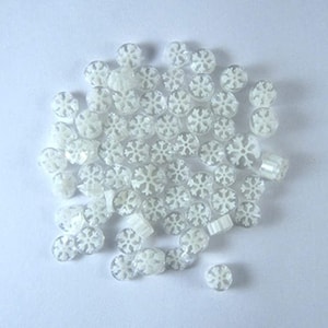 May include: A collection of clear, snowflake-shaped beads. The beads are white and have a detailed snowflake design. The beads are scattered on a white surface, creating a winter or holiday theme.