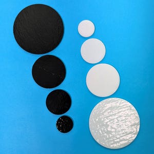 May include: An assortment of black and white round cardboard cake boards of varying sizes, arranged on a bright blue surface. The black boards have a textured, shiny finish, while the white ones have a smooth appearance. The sizes range from small to large.