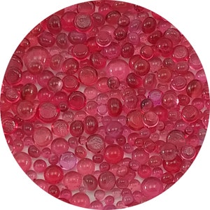 May include: A close-up view of a large quantity of small, round, translucent pink glass beads. The beads are scattered across a white surface.