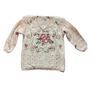 May include: A light pink sweater with long sleeves. The sweater features a floral design in the center, with pink and green accents. The neckline and shoulders are adorned with small, decorative embellishments. The sweater has a textured knit pattern.