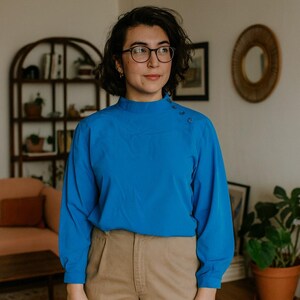 May include: A vibrant blue long-sleeved blouse with a high neckline and three small buttons on the shoulder. The blouse is worn with beige trousers. The person in the image is wearing spectacles.