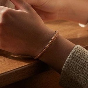 May include: A delicate rose gold bracelet with a textured surface. The bracelet is worn on a wrist, with a gray sweater sleeve visible. The background is a wooden surface.