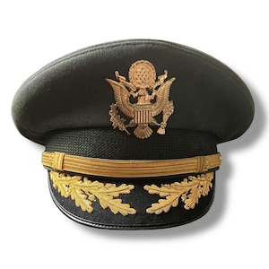 Może przedstawiać: Ciemnozielona czapka wojskowa ze złotym emblematem przedstawiającym orła i okrągły wzór. Czapka ma złoty pasek i złote akcenty liści na rondzie. Czapka jest w stylu wojskowym.