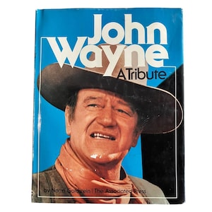 May include: A book cover with a portrait of John Wayne wearing a cowboy hat. The title "John Wayne A Tribute" is in white and tan lettering against a blue backdrop. The author and publisher are at the bottom.