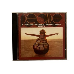 Puede incluir: Una caja de CD con la palabra "Decade" en rojo. La portada muestra a una persona con los brazos extendidos, su cuerpo adornado con portadas de álbumes, sobre un fondo desértico. El texto dice "A 3-Record Set on 2 Compact Discs Disc 1."