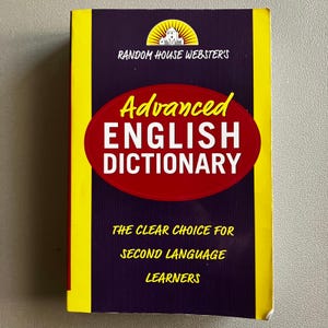 May include: A Random House Webster's Advanced English Dictionary. The book has a purple cover with yellow edges and a red oval with white text that reads "Advanced English Dictionary". The text "The Clear Choice for Second Language Learners" is also visible.