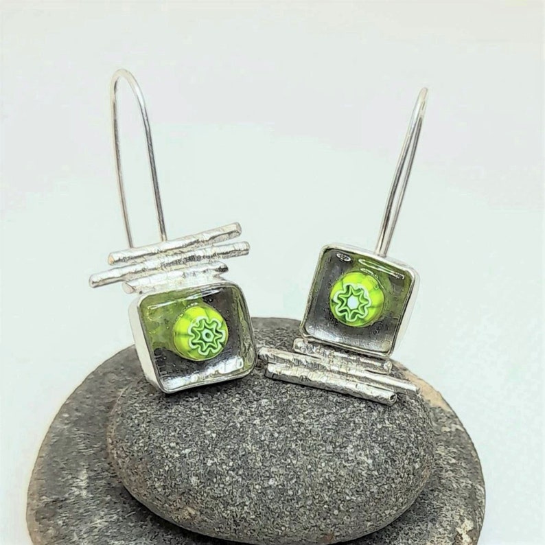 Pu&ograve; includere: Un paio di orecchini pendenti in argento con quadrati di vetro verde e bianco. Gli orecchini presentano un design geometrico con sottili barre d'argento.