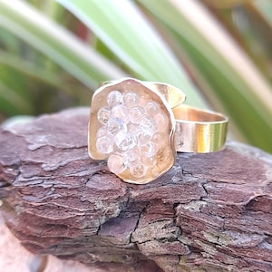 Op de afbeelding: Een goudkleurige ring met een uniek ontwerp. De ring heeft een grote, onregelmatig gevormde zetting gevuld met heldere, belachtige versieringen. De band is een eenvoudig, gepolijst goud. De ring wordt getoond op een stuk getextureerde, bruine schors.