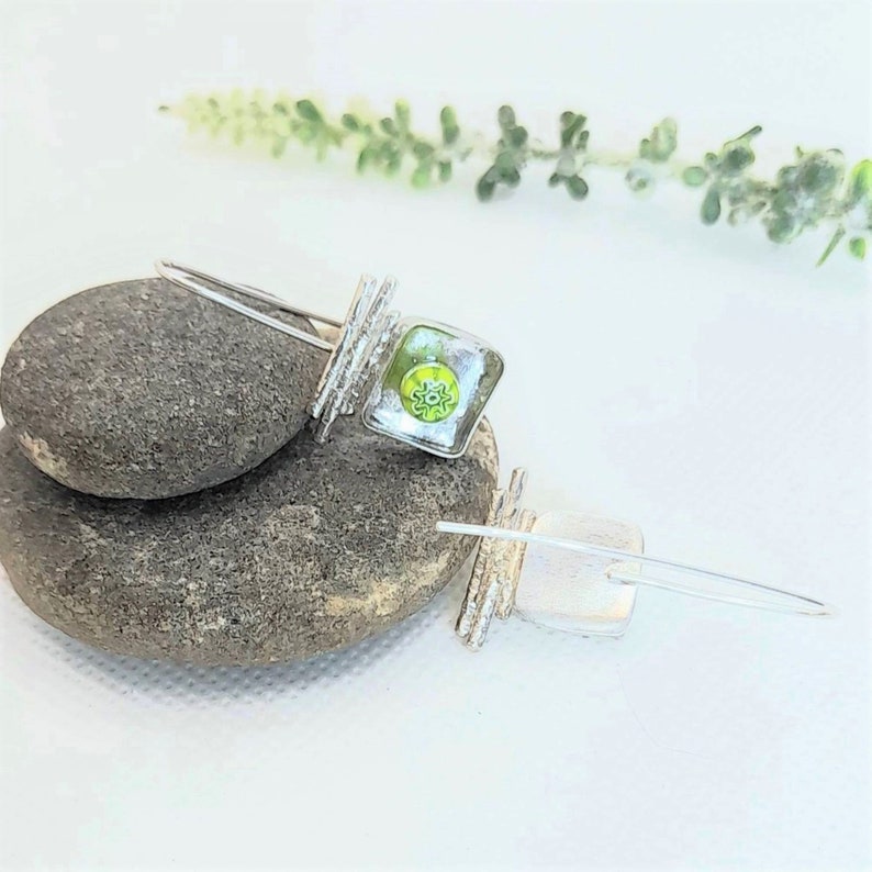 Pu&ograve; includere: Un paio di orecchini pendenti in argento con quadrati di vetro verde e trasparente. Gli orecchini sono su una superficie di pietra grigia.