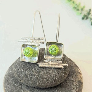 Pu&ograve; includere: Un paio di orecchini pendenti in argento con quadrati di vetro verde e bianco. Gli orecchini presentano un design geometrico con barre in argento.