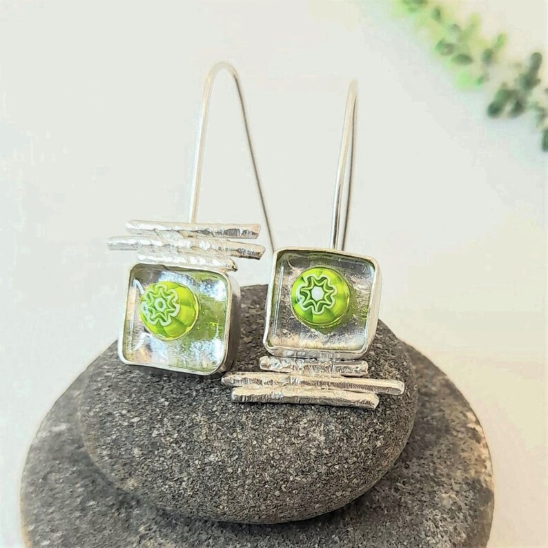 Pu&ograve; includere: Un paio di orecchini pendenti in argento con quadrati di vetro verde e bianco. Gli orecchini presentano un design geometrico con barre in argento.