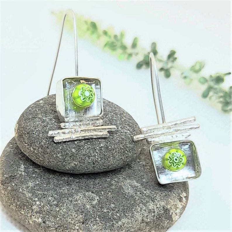 Pu&ograve; includere: Un paio di orecchini pendenti in argento con quadrati di vetro verde che presentano un motivo a fiore bianco. Gli orecchini sono appesi a una pila di pietre grigie.