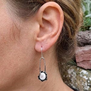 Op de afbeelding: Zilveren oorbel met een wit schelpmotief. De oorbel heeft een lange, slanke, gebogen zilveren haak die verbinding maakt met een decoratief zilveren frame. Het frame omringt een kleine, witte schelp met een spiraalpatroon.