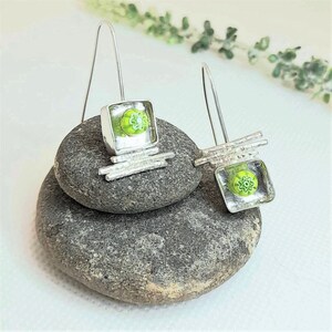 Pu&ograve; includere: Un paio di orecchini pendenti in argento con quadrati di vetro verde che presentano un motivo a fiore bianco. Gli orecchini sono esposti su una pila di pietre grigie.