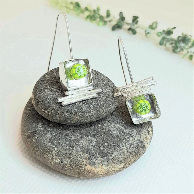 Pu&ograve; includere: Un paio di orecchini pendenti in argento con quadrati di vetro verde che presentano un motivo a fiore bianco. Gli orecchini sono esposti su una pila di pietre grigie.