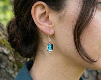 May include: A silver earring with a rectangular, light blue gemstone. The gemstone is set in a dark frame and hangs from a silver hook. A small silver ball is at the bottom of the earring.