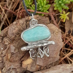 May include: A pendant featuring a smooth, oval-shaped light blue-green stone set in a textured, silver-toned metal frame. The metalwork includes a horizontal bar and three small, irregular, dark metallic accents hanging from the bottom.