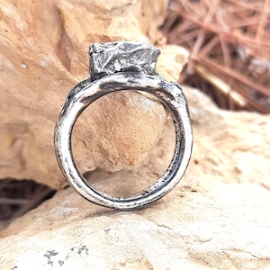 May include: A handcrafted silver ring with a textured band and a large, irregularly shaped stone setting. The ring has a rustic, organic design, showcasing a unique, artistic style. The ring is displayed on a natural, light-colored stone surface.