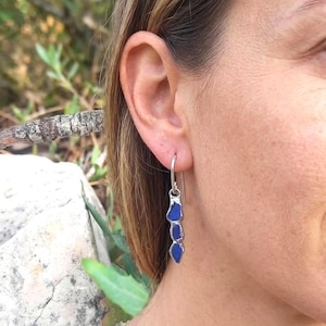 Può includere: Orecchini pendenti in argento con tre pezzi di vetro blu vibrante di forma irregolare. Gli orecchini sono sospesi a un semplice gancio. Il vetro blu ha l'aspetto del vetro marino.