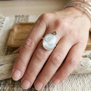 Può includere: Un anello d'argento con una pietra ovale bianca con un motivo a spirale. Una piccola pietra rotonda e marrone è incastonata sopra la pietra ovale. L'anello è indossato su un dito.