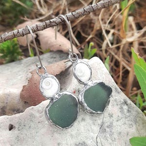 May include: Handmade earrings with sea glass and shell details. The earrings have a silver-coloured metal frame. The sea glass is a deep green, and the shells are white. The earrings are suspended from a branch, showcasing their unique design.