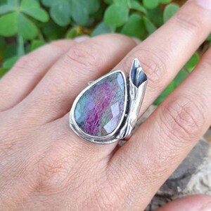 Op de afbeelding: Zilveren ring met een grote, peervormige edelsteen. De edelsteen heeft een facetgeslepen oppervlak en vertoont paarse, roze en groene tinten. De ring heeft een uniek, sculpturaal ontwerp met een getextureerd, organisch element aan de zijkant.
