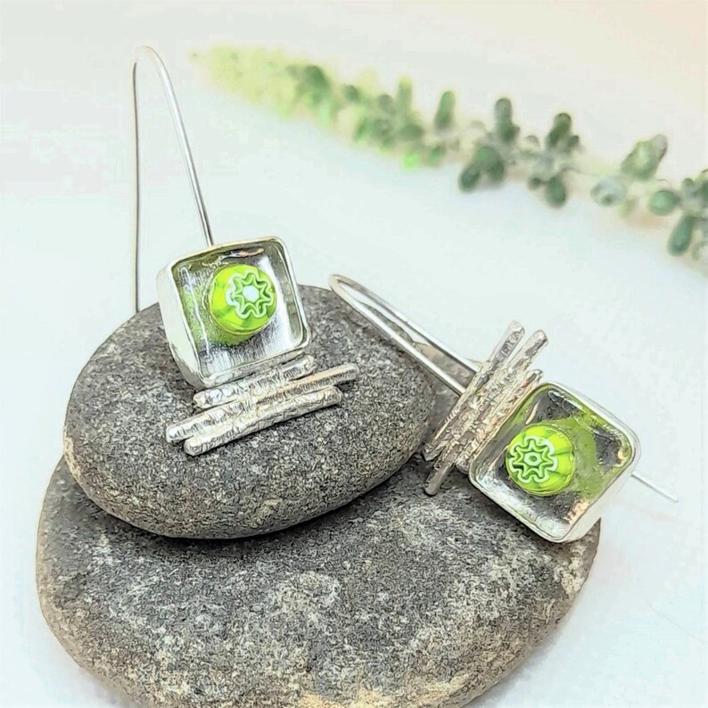Pu&ograve; includere: Un paio di orecchini d'argento con quadrati di vetro verde e un motivo a fiore bianco. Gli orecchini sono appesi a un filo d'argento e hanno un design a barra d'argento testurizzato.