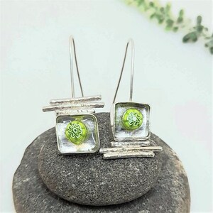 Pu&ograve; includere: Orecchini pendenti in argento con quadrati di vetro verde e barre argentate. I quadrati di vetro hanno un motivo a fiore bianco.