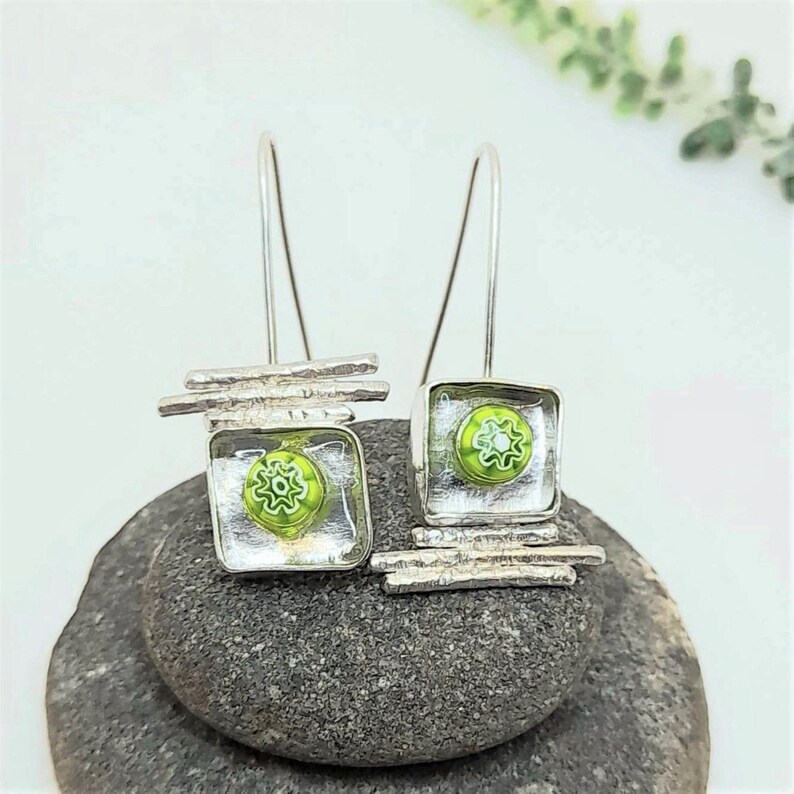 Pu&ograve; includere: Orecchini pendenti in argento con quadrati di vetro verde e barre argentate. I quadrati di vetro hanno un motivo a fiore bianco.