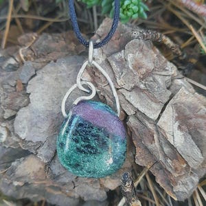 Puede incluir: Collar con colgante artesanal con una piedra preciosa ovalada pulida con tonos verdes, morados y negros. La piedra está suspendida de una fianza de metal plateado abstracto y un cordón negro. Joyería.
