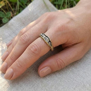 Op de afbeelding: Een goudkleurige ring met een uniek ontwerp, gezet met vijf heldere, onregelmatig gevormde stenen in een gebogen band. De ring wordt op een vinger gedragen, waarbij de organische, getextureerde uitstraling wordt benadrukt.