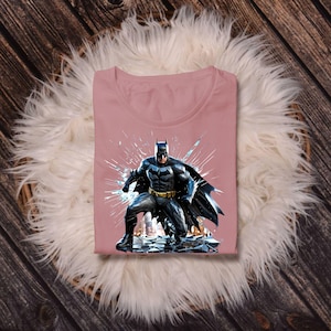 May include: A dusty rose t-shirt featuring a graphic of Batman in a dynamic pose. The superhero is depicted in a black and gray suit with a flowing cape, set against a backdrop of shattered glass. The shirt is folded neatly.