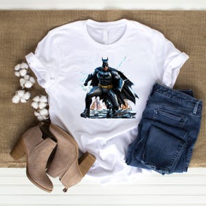 May include: White t-shirt featuring a graphic of Batman in a dynamic pose. The superhero is depicted in a black and gray suit with a flowing cape. The shirt is paired with blue jeans and beige heeled boots, suggesting a casual, everyday style.