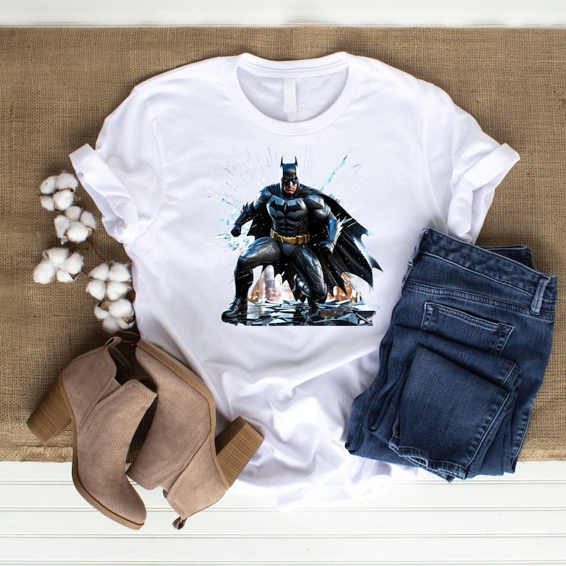 May include: White t-shirt featuring a graphic of Batman in a dynamic pose. The superhero is depicted in a black and gray suit with a flowing cape. The shirt is paired with blue jeans and beige heeled boots, suggesting a casual, everyday style.