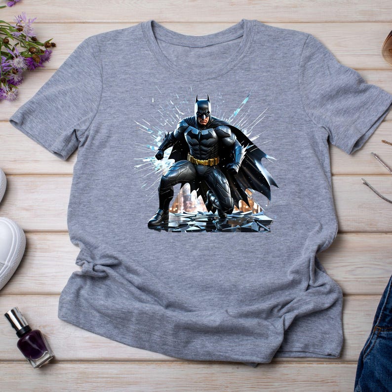 May include: A gray t-shirt featuring a detailed graphic of Batman in a dynamic pose. The superhero is depicted in his iconic black and gray suit, with a yellow utility belt, set against a shattered glass effect. The shirt is made of a soft material.