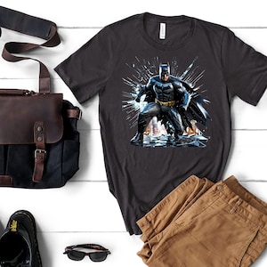 May include: Dark gray t-shirt featuring a graphic of Batman in a dynamic pose. The superhero is depicted in a black and gray suit with a yellow belt, set against a backdrop of shattered glass. Tan pants, a black bag, and sunglasses are also visible.