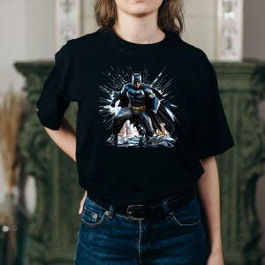 May include: Black t-shirt featuring a graphic of Batman in a dynamic pose, with a dark cape and a yellow utility belt. The artwork has a comic book style with a shattered glass effect in the background.