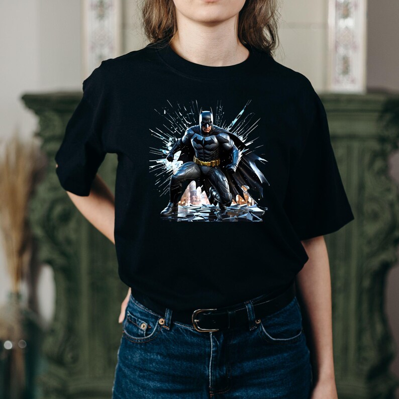 May include: Black t-shirt featuring a graphic of Batman in a dynamic pose, with a dark cape and a yellow utility belt. The artwork has a comic book style with a shattered glass effect in the background.
