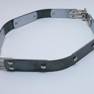 Könnte beinhalten: Ein silberner Gürtel aus Metall mit einer Scharnier-Schnalle und Nieten. Der Gürtel besteht aus mehreren Abschnitten, die durch die Nieten verbunden sind. Der Gürtel ist dunkelgrau.