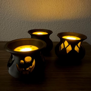 May include: Three black candle holders with cut-out designs, each containing a glowing yellow light. The front holder features a skull-like face pattern, while the others have diamond-shaped cut-outs, all emitting a warm, bright illumination.